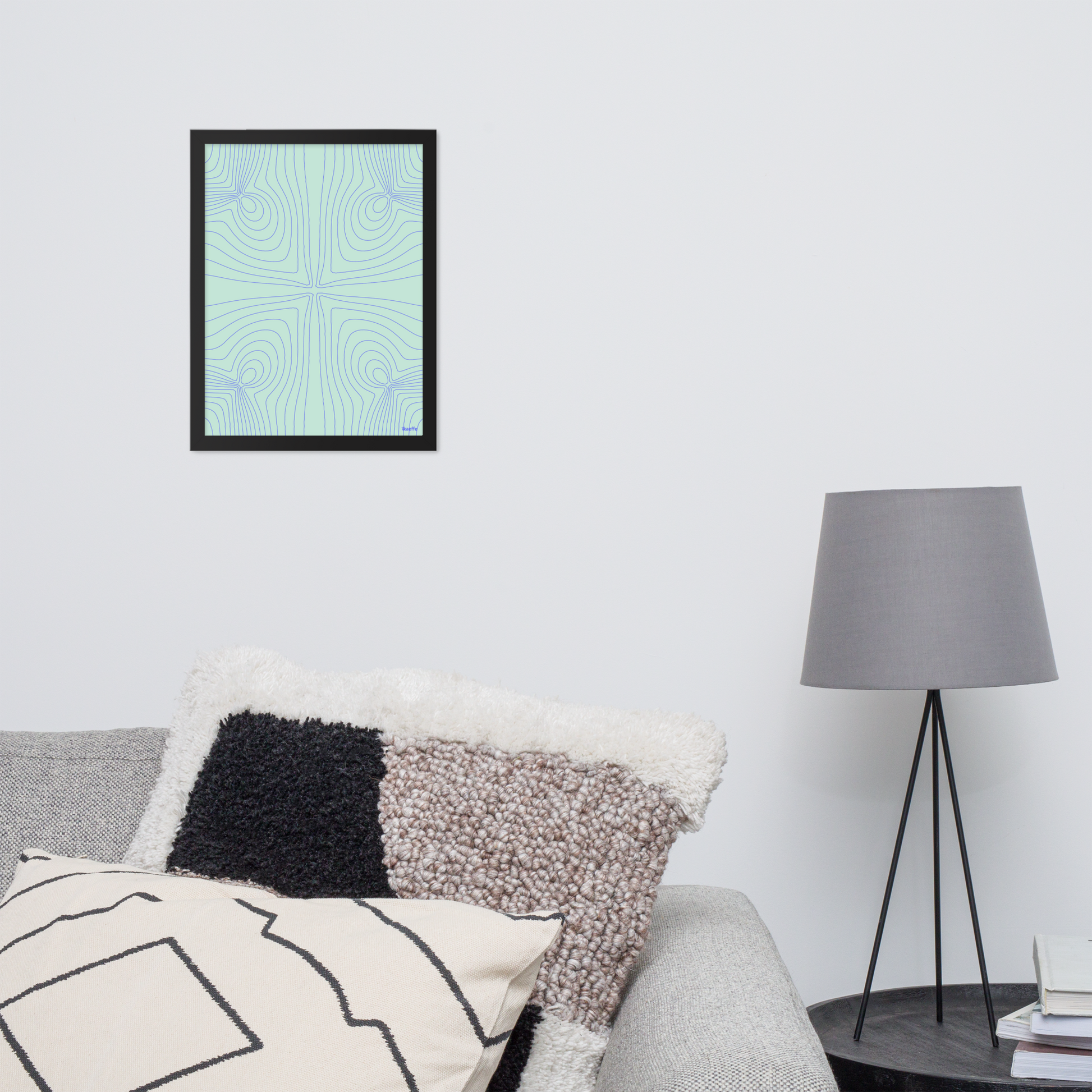 Ein minimalistischer Raum mit einer grauen Stehlampe und einem grau-weißen Sofa mit einem gemusterten Kissen. Auf dem Sofa liegt eine Decke mit verschiedenen Texturen. Ein gerahmtes Kunstwerk, Kunstdruck – θ von Kaeffe Art, mit blauen und grünen Wellenlinien hängt in einem mattschwarzen Holzrahmen an der weißen Wand über dem Sofa.