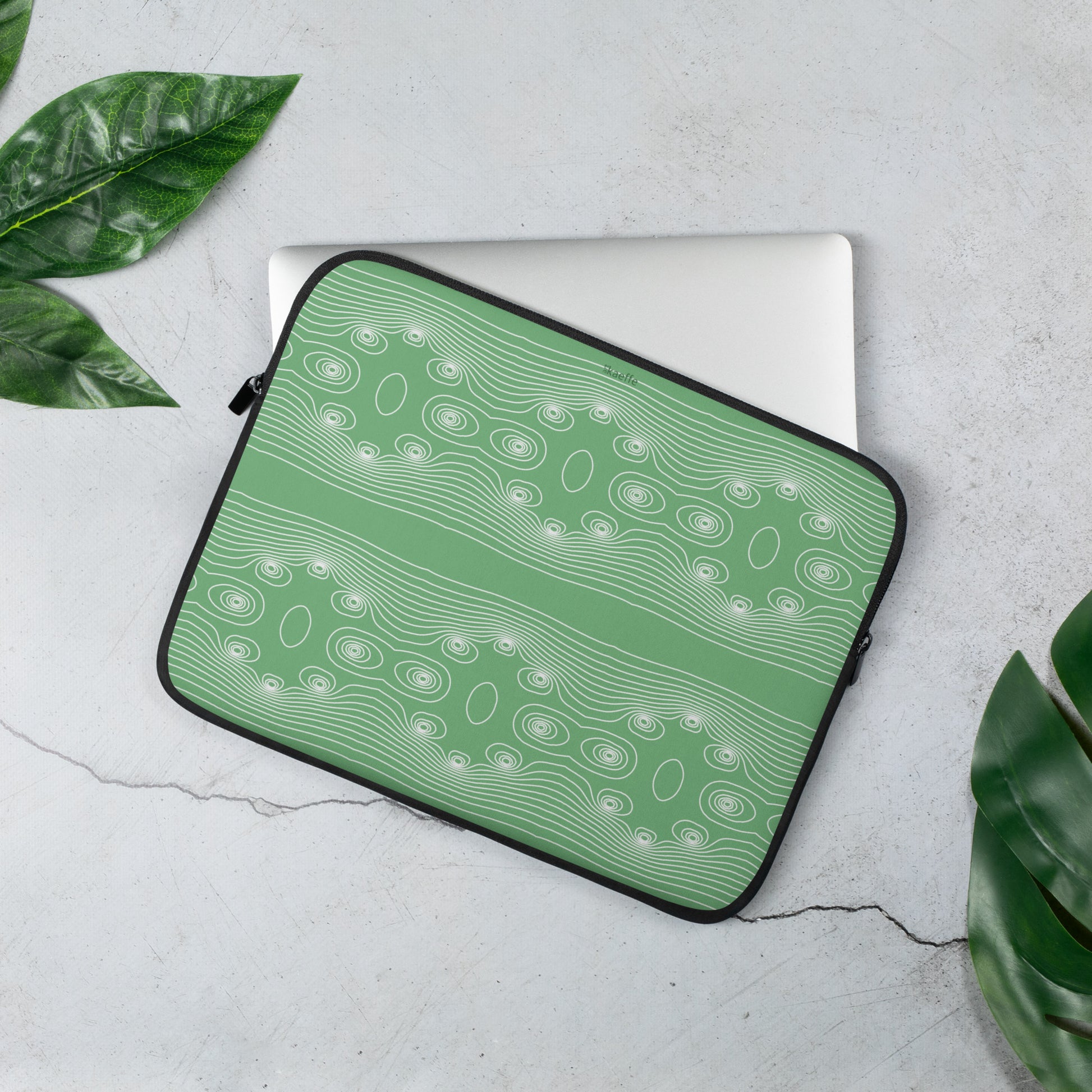 Eine grüne Kaeffe Art Laptoptasche - ε, verziert mit einem weißen Wellenlinienmuster, liegt teilweise offen auf einer grauen, rissigen Betonoberfläche. Die umgebenden grünen Blätter verstärken die Atmosphäre und bringen ein natürliches Element ein. Die Hülle ist aus wasserfestes Material gefertigt und verfügt über Kunstfell-Innenfutter für zusätzlichen Schutz. Im Inneren ist ein teilweise sichtbarer Laptop zu sehen.